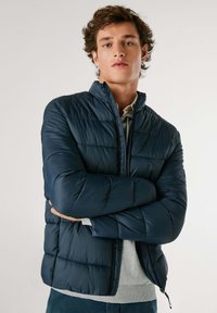 Mornarsko puhovko z zadrgo spredaj, vodoravnimi šivi in stojnim ovratnikom. Oblečeno čez svetlo sivo majico, ki prikazuje gladko teksturo.