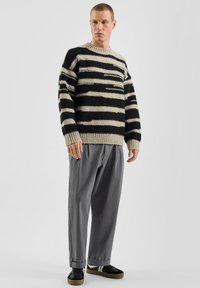 Pull en maille épaisse à rayures noires et beiges, col rond, poignets et ourlet côtelés. Pantalons gris ample et baskets noires.