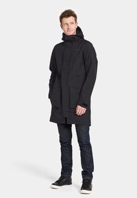 Sort vandtæt parka med hætte, to frontlommer og velcro-manchetter, parret med mørke jeans og sorte sneakers. Glat tekstur.