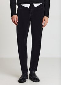 Schwarze Cordhose mit geradem Schnitt, feiner Rippstruktur, Fronttaschen und kombiniert mit einem weißen Hemd unter einem schwarzen Pullover.