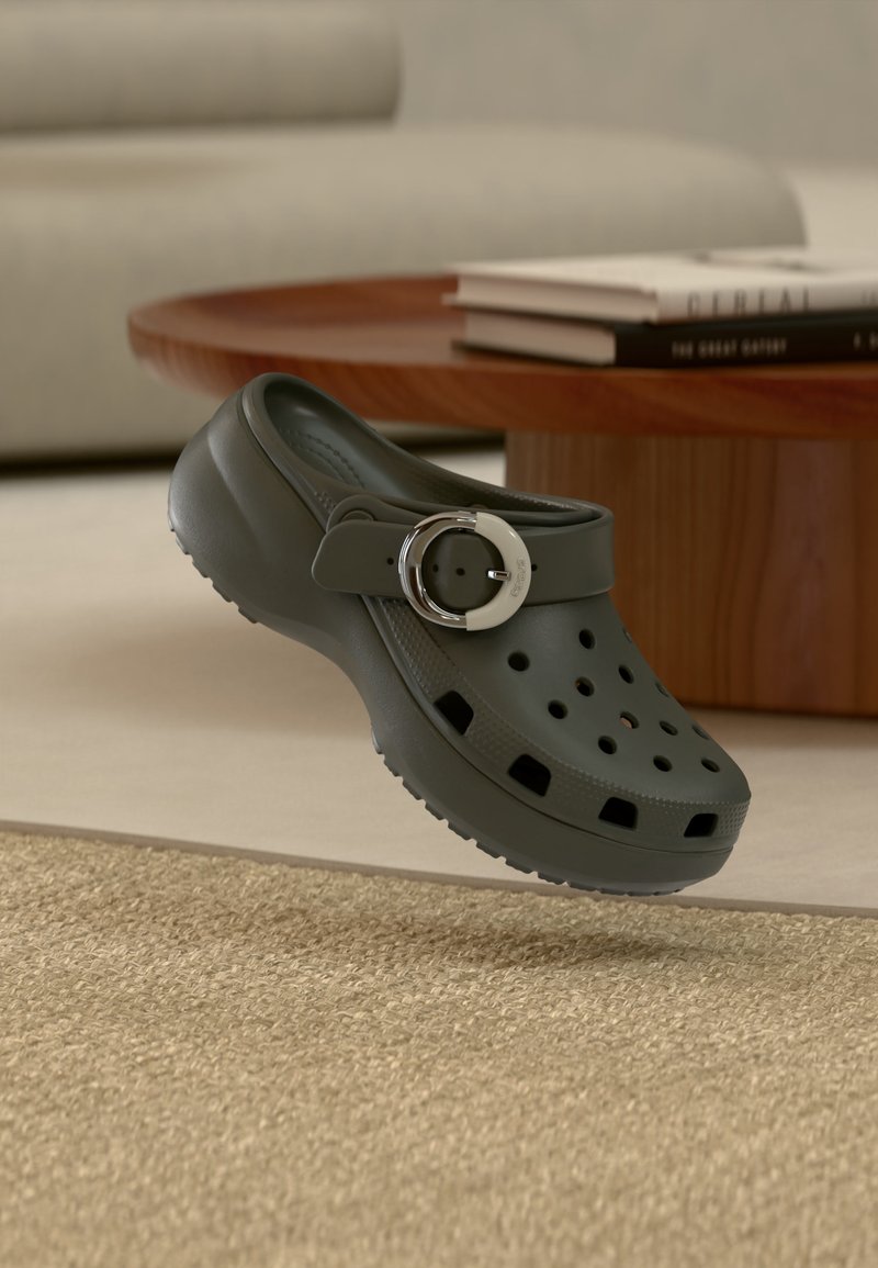 Σκούρο γκρι παπούτσι τύπου κλογκ με οπές για εξαερισμό και ασημένιο λουράκι με πόρπη, τοποθετημένο πάνω σε μπεζ χαλί σε περιβάλλον σαλονιού.