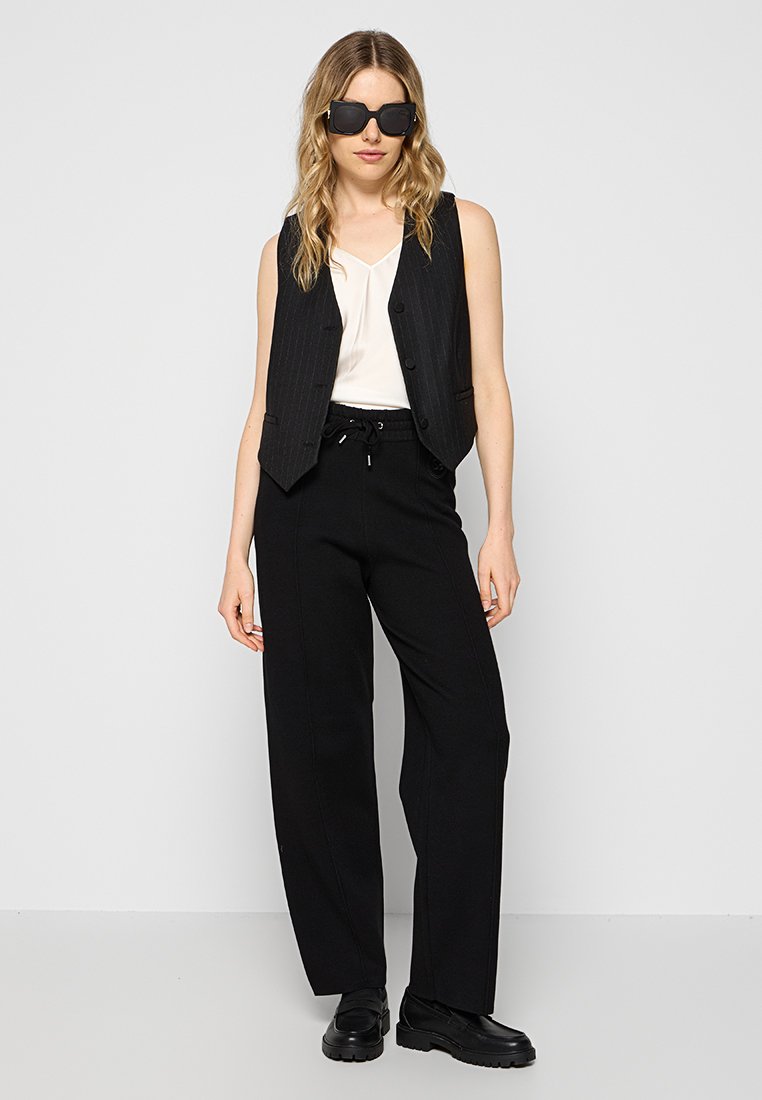 Schwarze Streifenweste über einer weißen Bluse, kombiniert mit lockeren schwarzen Hosen. Model trägt schwarze Sonnenbrillen und klobige schwarze Schuhe.