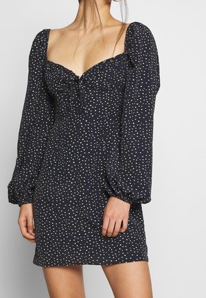 Abito mini blu navy con lunghe maniche a palloncino, scollo quadrato e pois gialli. Presenta un corpetto aderente e un laccetto nella parte anteriore.
