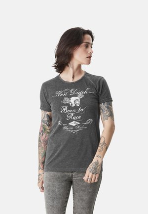 Jeune personne aux cheveux foncés mi-longs et tatouages sur les bras, portant un t-shirt gris "Von Dutch Born to Race" et un jean gris, regardant vers la droite.