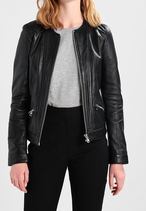 Frau trägt eine schwarze Lederjacke mit Reißverschluss über einem hellgrauen Shirt und schwarzen Hosen, steht vor einem schlichten Hintergrund.