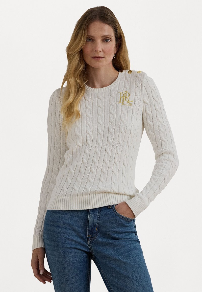 Bela kablasta pletena pulover s okruglim izrezom, rebrastim manžetama i rubom, s zlatnim grbom na prsima i dekorativnim gumbima na ramenu.