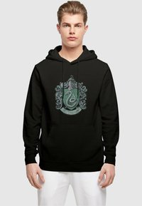 Svart hettegenser med Slytherin-våpen i grønne og sølvfargede detaljer, inkludert en detaljert slange og dekorative elementer. Kengurulomme foran.