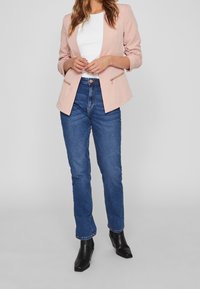 Blazer rose poudré à coupe ajustée, avec poches à fermeture éclair, associé à un jean bleu taille haute et des bottines noires.