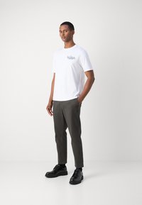 Valkoinen lyhythihainen t-paita, jossa on musta graafinen kuvio vasemmassa rinnassa, yhdistettynä harmaisiin slim fit -farkkuihin ja mustiin nauhakenkiin.