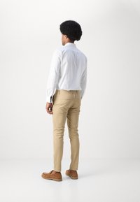 Witte button-down shirt, tan slim-fit broek, bruine suède instapschoenen met rubberen zolen; gefotografeerd vanachter tegen een effen achtergrond.