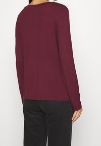 Burgunderfarbene Langarmbluse in lässiger Passform, mit weichem Material und rundem Ausschnitt. Kombiniert mit schwarzen Hosen, was einen lässigen Look erzeugt.