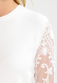 Weißes, geripptes Oberteil mit durchsichtigen Spitzenlangärmeln und floralen Stickdetails. Glatte Textur mit feinen Mustern an den Ärmeln.