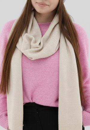 Beige knitted scarf drapes loosely over a pink sweater. The scarf features a textured pattern and fringed ends. Soft materials emphasize comfort.