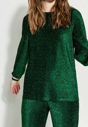 Osoba ubrana w błyszczącą zieloną bluzkę z długim rękawem i dopasowane spodnie, stojąca na tle jasnego, jednolitego tła.