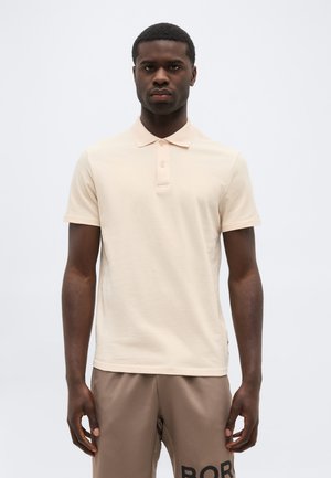 Férfi bézs rövid ujjú pólóingben és barna rövidnadrágban, fekete felirattal, egy egyszerű fehér háttér előtt állva.