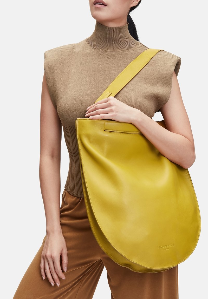 Une femme portant un haut beige sans manches et un pantalon marron tient un grand sac à bandoulière en cuir jaune d'une main devant sa poitrine.