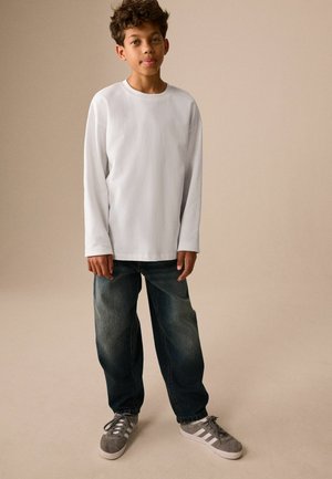 Dreng med krøllet hår iført en hvid langærmet skjorte, løse mørke jeans og grå sneakers, der står mod en ensartet beige baggrund.