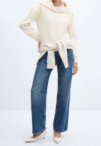 Pull en maille couleur crème avec capuche, noué à la taille par-dessus un jean taille haute bleu clair et des talons blancs pointus. Texture lisse.