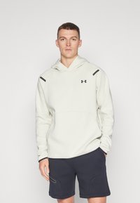 Hellebeige Kapuzenpullover mit einer Fronttasche, schwarzen Akzenten an den Schultern und einem Under Armour-Logo. Kombiniert mit dunkelblauen Shorts mit seitlichen Details.