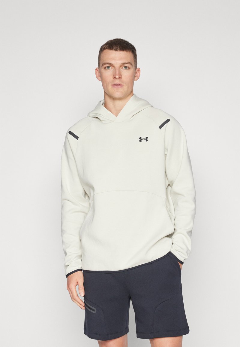 Hellebeige Kapuzenpullover mit einer Fronttasche, schwarzen Akzenten an den Schultern und einem Under Armour-Logo. Kombiniert mit dunkelblauen Shorts mit seitlichen Details.