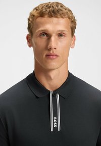 Sort polo shirt med klassisk krave, knaplukning og en mærket lodret stribe i hvid med "BOSS" tekst. Glat stofoverflade.