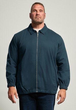Man met kort haar die een donkerblauwe jas met ritssluiting en elastische boorden draagt en staat tegen een effen lichte achtergrond.