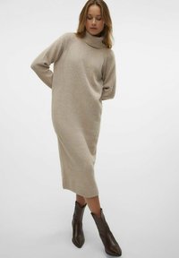 Kremna turtleneck obleka iz mehkega pletenega materiala, ohlapen kroj, dolgi rokavi, rebrasti detajli ter dolžina do sredine meč. Rjave gležnjarje.