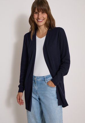 Femme souriante portant un cardigan bleu marine ouvert devant, un t-shirt blanc et un jean bleu clair, une main dans la poche, sur un fond uni.
