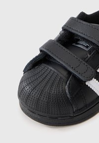 Baskets en cuir noir avec un capuchon de orteil texturé, fermeture à bande Velcro et rayures blanches. Dotées d'une semelle en caoutchouc avec un motif antidérapant.