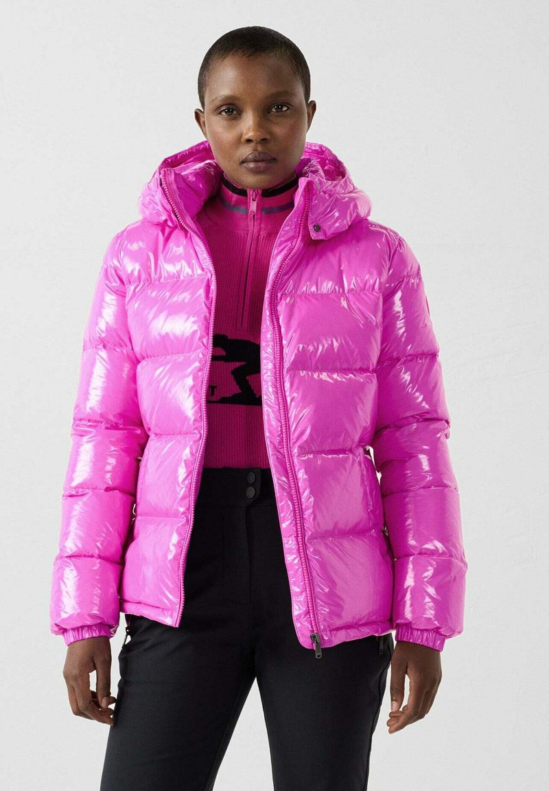 Giacca imbottita rosa lucida con cappuccio, design trapuntato e polsini a costine. Indossata sopra un maglione rosa aderente, abbinata a pantaloni neri.