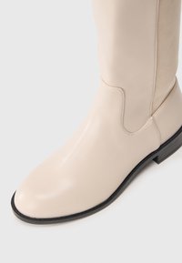 Beige læder støvle med knæhøjde, glat finish, rund tå, minimal syning og en lav sort hæl. Sidepanelet har en ruskindsstruktur.