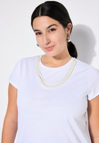 T-shirt bianco a maniche corte con tre collane di perle stratificate di diverse dimensioni, che creano un aspetto elegante e textures.