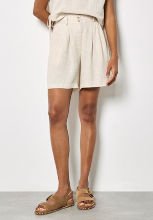 Persoon met beige, plissé hoge taille shorts en tan open-toe sandalen, staand tegen een witte muur en een grijze vloer.