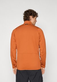 Oransje langermet jakke med høy krage, strukturert stoff, baklomme og svart logo plassert øverst på ryggen.