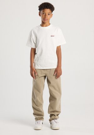 Junge steht da und trägt ein weißes T-Shirt mit kleinem Logo, beige lockere Hose und weiße Sneaker vor einem schlichten hellen Hintergrund.