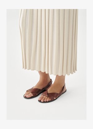 Rudos odinės basutės su kryžminėmis dirželiais, plokščiu padu ir atvirais pirštais, derinamos su kremine klostuota suknele.