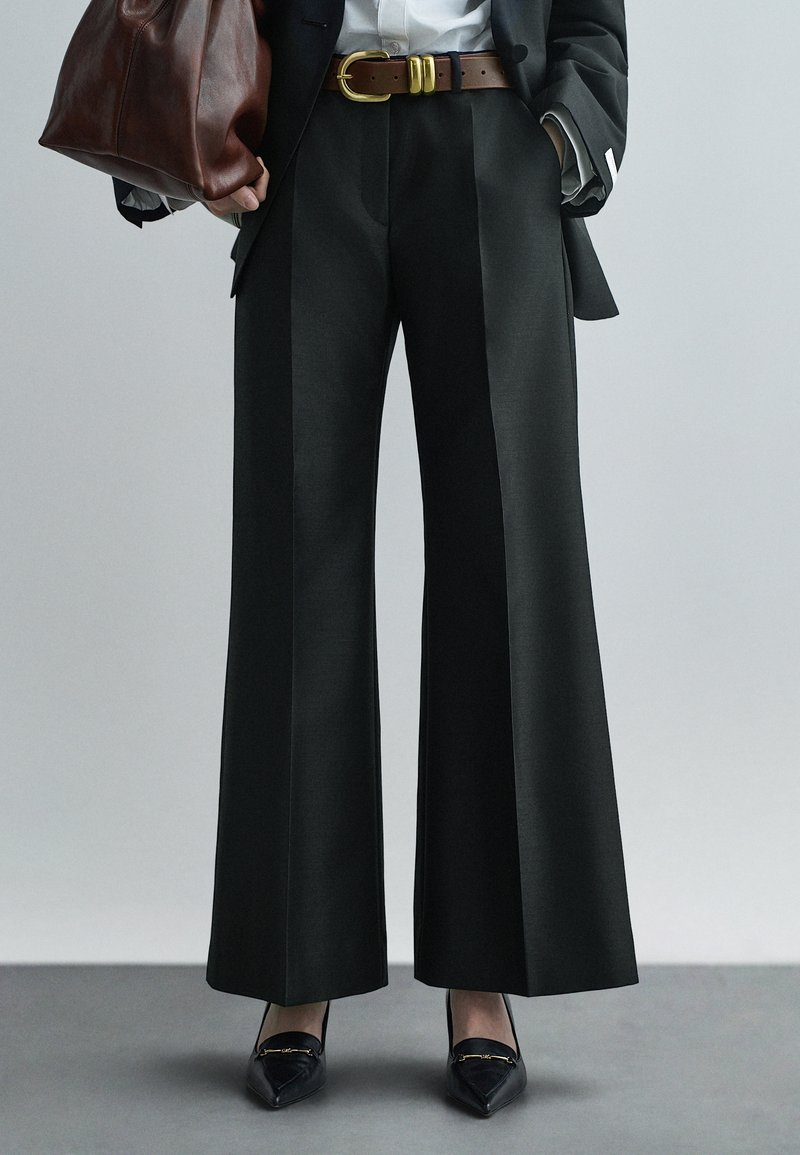 Wijdvallende donkere broek met een gladde textuur, plooidetails en gecombineerd met zwarte puntige schoenen; voorzien van een bruine riem met gouden gespsluiting.