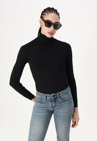 Svart genser med høy hals og lange ermer, parret med lyseblå jeans. Stoffet er glatt, tettsittende og elastisk, noe som fremhever en slank silhuett.