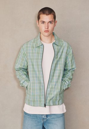 Ung mand med kort hår iført en grøn ternet jakke med lynlås over en hvid skjorte og blå jeans mod en ensfarvet beige baggrund.
