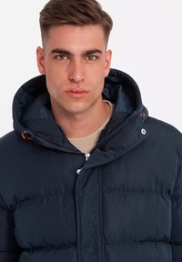Chaqueta acolchada azul marino con capucha, presenta un cierre con cremallera, botones a presión y detalles en naranja. Textura suave y diseño acolchado.