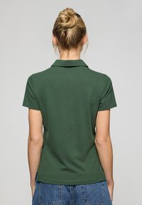 Zielona koszulka polo z kołnierzykiem, krótkim rękawem i gładką teksturą. Pokazana z tyłu, noszona z szortami dżinsowymi o wysokim stanie.