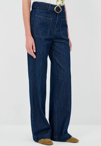 Jeans a vita alta di colore blu scuro, a gamba larga, con una cintura dotata di una fibbia circolare oro, due tasche posteriori e dettagli di cuciture a contrasto.