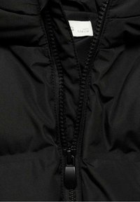 Gros plan sur une veste matelassée noire avec une fermeture éclair partiellement ouverte et une étiquette de taille blanche indiquant "128 CM" dans la zone du col.