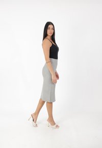 Zwarte mouwloze top, grijze getailleerde rok tot op de knie en witte open hakken met gouden accenten. Model poseert in profiel tegen een effen achtergrond.