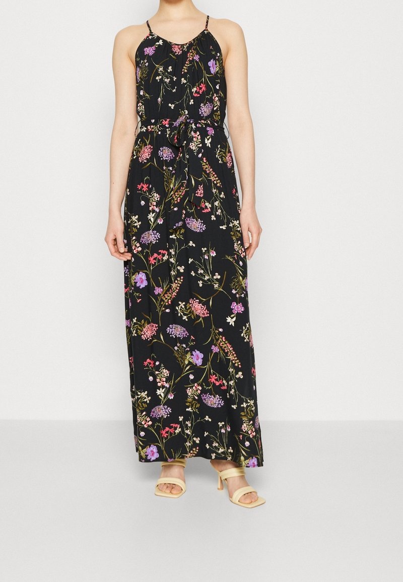 Femme portant une robe maxi noire à fleurs avec des bretelles fines et une ceinture à la taille, associée à des sandales à talons beiges.