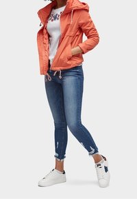 Orange lättviktsjacka med dragsko, kombinerad med en vit blommig t-shirt, slitna blå jeans och vita joggingskor.