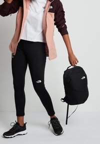 Black backpack with The North Face logo, worn with a pink and maroon jacket, black leggings, and black sport shoes. White t-shirt visible.