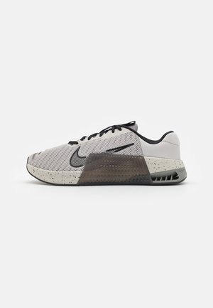 Chaussure de sport gris clair avec des lacets noirs, une semelle intermédiaire texturée avec des taches noires, un logo Nike noir et un soutien de talon transparent.
