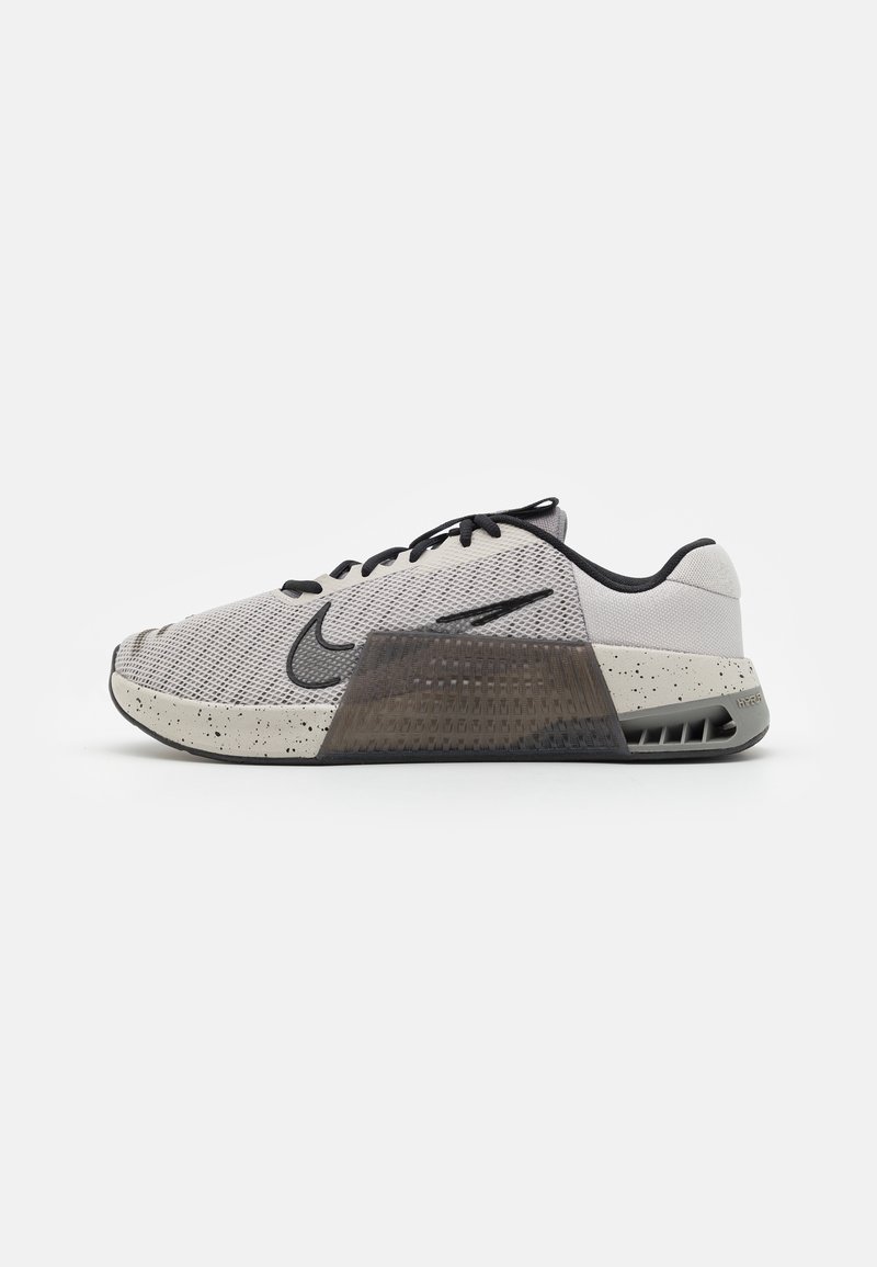 Chaussure de sport gris clair avec des lacets noirs, une semelle intermédiaire texturée avec des taches noires, un logo Nike noir et un soutien de talon transparent.
