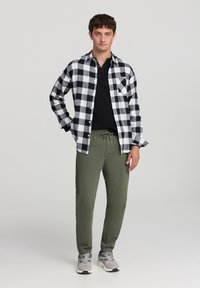 Koszula z flaneli w kratkę w czarno-białym kolorze, nałożona na czarną bluzkę, w komplecie z oliwkowymi joggerami i szarymi butami sportowymi. Luźny krój.